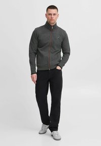 Sivi zip-up jakna s visokim ovratnikom, s kontrastnim narančastim zatvaračem i šavovima, u kombinaciji s crnim hlačama i sivim tenisicama.