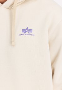 Beige kapuce ar mīkstu audumu un kenguru kabatu, ar purpura "Alpha Industries" logotipu uz krūšu un aukliņu kapuci.