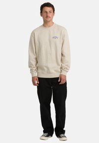 Beżowa bawełniana bluza z ściągaczami przy mankietach i dolnej krawędzi, z jasnoniebieskim graficznym logo na piersi. W zestawie z czarnymi spodniami i butami.