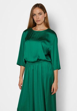 Woman with long brown hair wearing a shiny green satin dress with 3/4 sleeves and a gathered waist, standing against a plain white background.