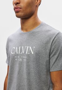 Zbliżenie na osobę w szarym T-shircie z białym napisem "CALVIN NEW YORK 1968 CK Calvin Klein" na klatce piersiowej.