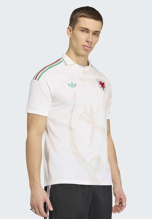 Joven con camiseta deportiva blanca con emblema de dragón rojo y logo verde de Adidas, posando frente a un fondo claro y sencillo.