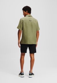 Maglietta verde oliva a maniche corte in lino con taglio dritto, dettaglio del tag sul retro, abbinata a pantaloni corti neri e scarpe da ginnastica nere.