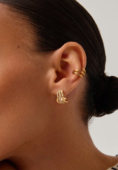 Χρυσά σκουλαρίκια στο αυτί μιας γυναίκας: ένα μικρό στριφογυριστό στεφανάκι στο πάνω μέρος του αυτιού και ένα κυματιστό, υφασμένο καρφάκι στο λοβό του αυτιού.