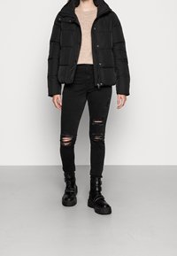 Sort puffet jakke med høj krave, beige sweater, sorte ripped jeans og kraftige sorte støvler med spænder og strukturerede såler.