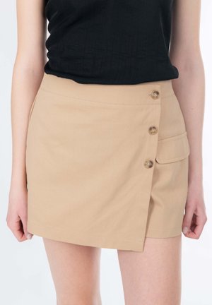 Mini-jupe beige en tissu lisse, avec des boutons latéraux et un détail poche. Portée avec un top noir texturé.