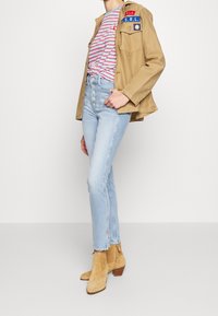 Person wearing tan jacket with patches, striped red and white shirt, high-waisted light blue jeans, and tan suede ankle boots.