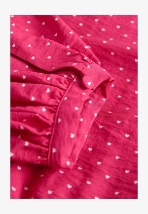 Rózsaszín anyag apró, fehér szívmintákkal, összegyűrt mandzsettával és gombos részlettel. Puha textúrájú és könnyű kialakítású.