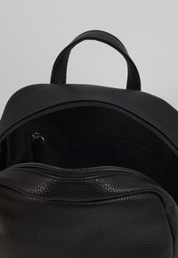 Sac à dos en cuir noir avec une surface texturée, doté d'une fermeture éclair et d'une petite poche intérieure, au design minimaliste, avec une poignée supérieure.