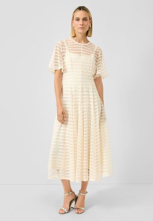 Frau in einem cremefarbenen, transparent gestreiften Midikleid mit flatternden Ärmeln und silbernen High-Heel-Sandalen, die vor einem schlichten Hintergrund steht.