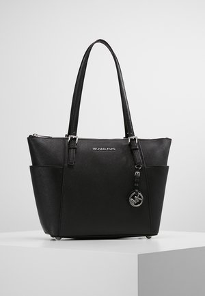 Sac cabas noir Michael Kors avec doubles poignées, poches latérales et un pendentif logo argenté suspendu près de la fermeture éclair du dessus.