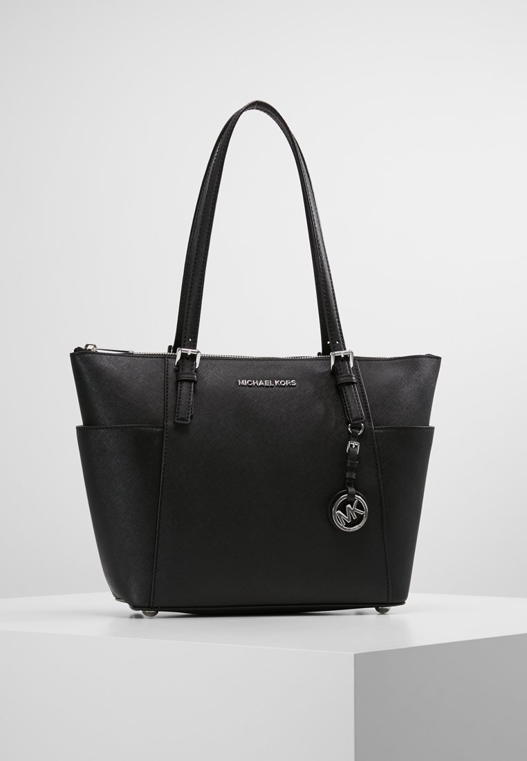 Sac cabas noir Michael Kors avec doubles poignées, poches latérales et un pendentif logo argenté suspendu près de la fermeture éclair du dessus.