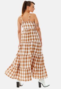 Maxi dress in brown and white checkered pattern, featuring a tiered design, spaghetti straps, and a flowing silhouette. Paired with white block heels.