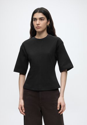 Ung kvinde med langt mørkt hår iført en sort kortærmet tætsiddende top og mørke bukser, stående mod en ensfarvet hvid baggrund.