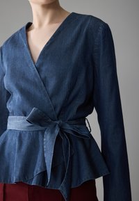 Blusa avvolgente in denim di un blu intenso, presenta una scollatura a V, orlo peplo e cintura in tessuto coordinato in vita. Texture morbida con maniche lunghe.