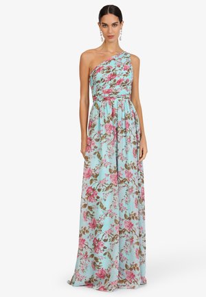 Frau trägt ein ärmelloses, einseitig schulterfreies, bodenlanges Kleid in Hellblau mit pinkem und braunem Blumenmuster, steht vor einem schlichten Hintergrund.