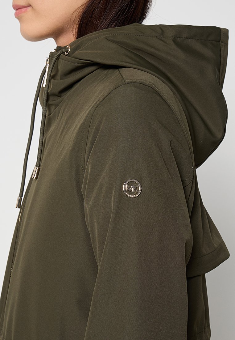 Kobieta w oliwkowym, zapinanym na zamek kapturze z metalowym logo na górnej części rękawa, widziana z boku.