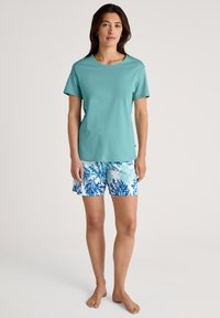 Light teal t-shirt with short sleeves paired with blue and white patterned shorts featuring coral designs. Barefoot model stands upright.