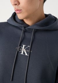 Temno modra bombažna jopica s kapuco, ki vključuje vrvico, vezeno logo "CK" in napis "Calvin Klein Jeans" na prsih. Gladka tekstura.