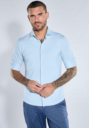 Mann mit kurzen Haaren und Tätowierungen auf den Unterarmen trägt ein hellblaues Hemd mit Knöpfen und hochgekrempelten Ärmeln sowie dunkelblaue Hose vor einem einfarbigen Hintergrund.