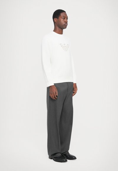 Sudadera blanca de algodón con un logo texturizado en el pecho, combinada con pantalones de pierna ancha grises y zapatos negros. Corte relajado, diseño minimalista.