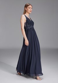 Robe de soirée bleu marine avec bodice en dentelle florale, décolleté en V et jupe fluide. Les accents semi-transparents et la longueur jusqu'aux chevilles accentuent l'élégance.