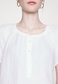 Blusa branca leve com mangas curtas, decote redondo e fecho frontal com pequenos botões combinando.