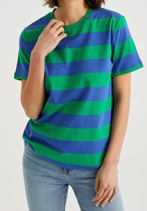 Kobieta ubrana w niebiesko-zielony szeroki poziomo paskowany t-shirt i jasnoniebieskie jeansy, z jedną ręką przy szyi, a drugą opartą z boku.