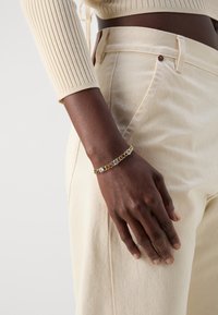 Gold chain bracelet featuring alternating clear stones, worn on a dark skin hand against a light-colored textured fabric background.