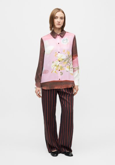 Žena oblečená v ružovo-hnedej kvetinovej košeli na gombíky s dlhým rukávom, čiernych a hnedých vertikálne pruhovaných širokých nohaviciach a čiernych topánkach.