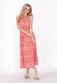 Rochie maxi roz cu gât halter și model geometric colorat; are un corset ajustat și o parte de Jos evazată, asortată cu sandale cu toc din beige.