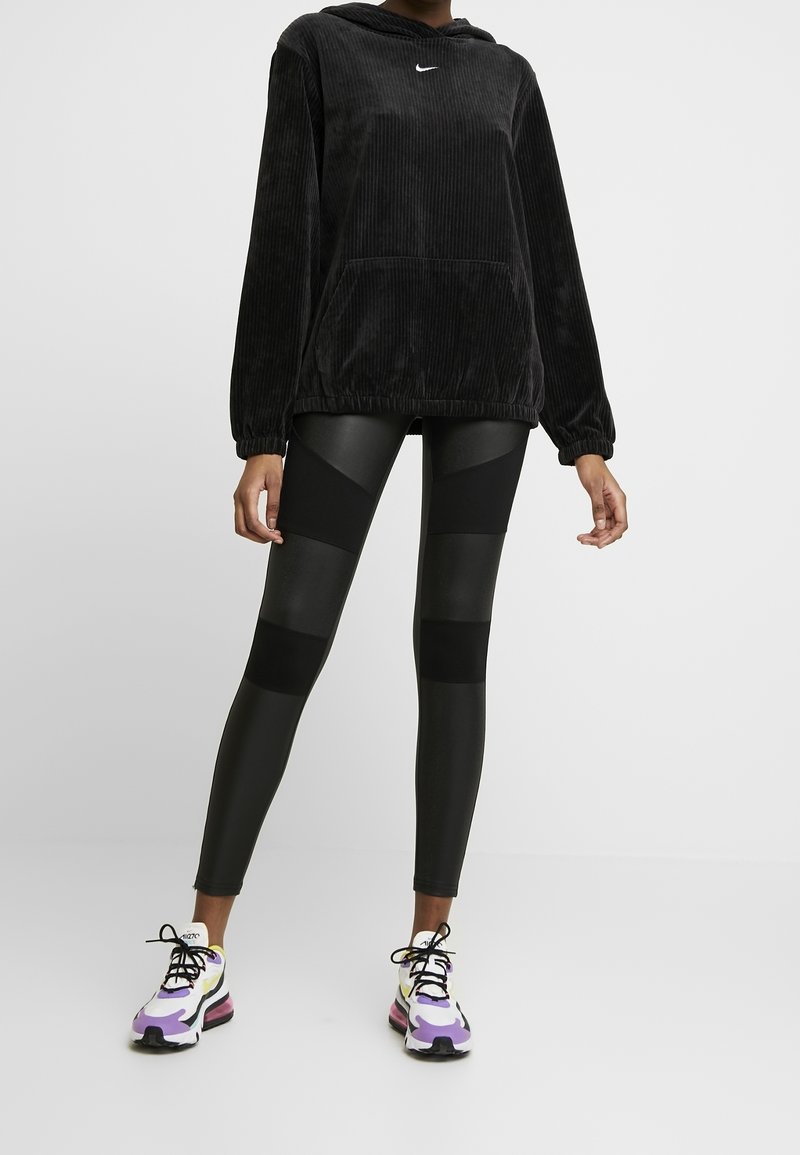 Zwarte corduroy hoodie met een voorzak en logo, gecombineerd met zwarte leggings met verschillende texturen en patronen, en multicolore sneakers.