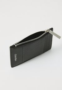 Portefeuille en cuir noir avec une finition texturée, fermeture éclair et étiquette en métal. Présente un logo Calvin Klein sur le côté et une forme rectangulaire élégante.