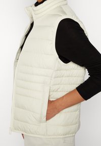 Gepolsterte ärmellose Weste in Off-White, mit stehendem Kragen, horizontalem Steppmuster und Seitentaschen. Das Material wirkt glatt und synthetisch.