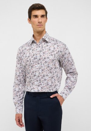 Mann trägt ein weißes langärmliges Hemd mit einem kleinen, mehrfarbigen Blumenmuster und dunkelblauen Hosen.