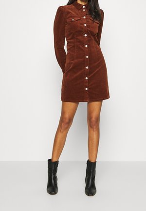 Braunes Cordkleid mit langen Ärmeln, Knopfleiste vorne, Kragen und zwei Brusttaschen; kombiniert mit schwarzen Knöchelstiefeln.