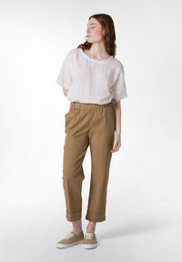 Camicia di lino bianca ampia, pantaloni con vita elastica marroni e tasche laterali, e sneakers in tela beige; il modello è in piedi con una leggera inclinazione.