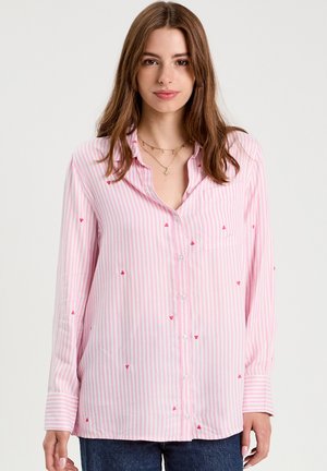 Young woman wearing a pink and white striped button-up shirt with small red heart patterns, paired with blue jeans, standing against a neutral background.