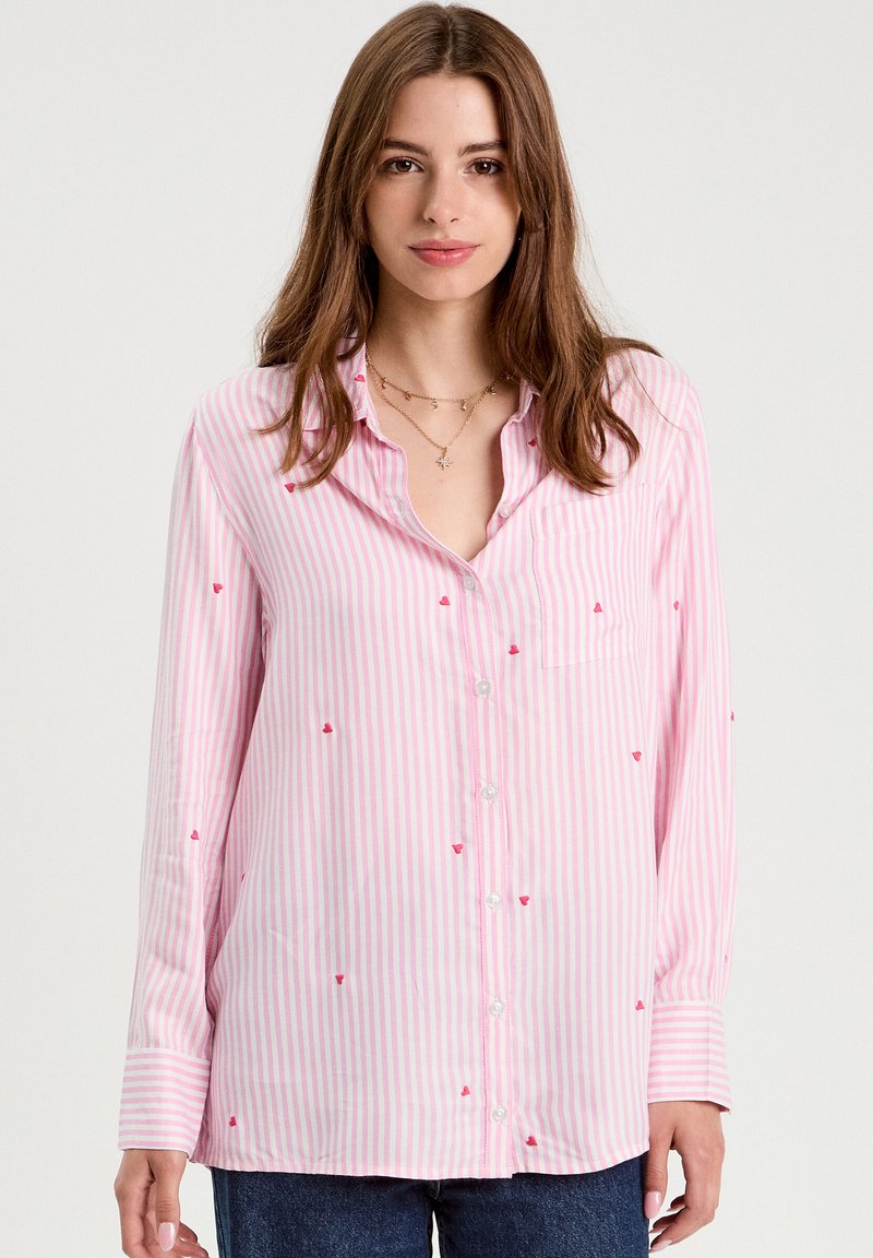 Joven con una camisa de botones de rayas rosas y blancas con pequeños patrones de corazones rojos, combinada con jeans azules, de pie frente a un fondo neutro.
