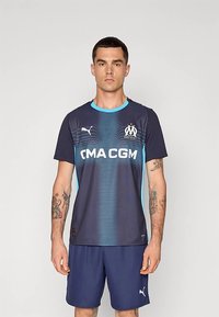 Tengerészkék sportpóló világoskék részletekkel és logóval, sima anyagból készült. Gradiens mintával és kerek nyakkivágással rendelkezik. Tengerészkék rövidnadrággal párosítva.