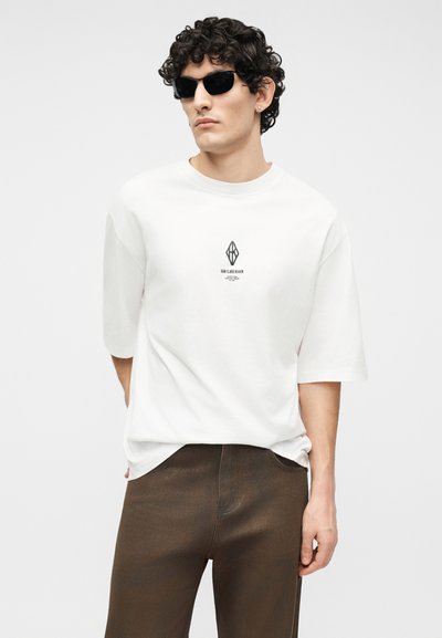 Göndör hajú férfi fekete napszemüveggel, fehér, túlméretezett pólóval, kis fekete logóval és barna nadrágban, egyszínű háttér előtt állva.