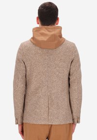 Uomo che indossa un blazer beige a scacchi con una sciarpa con cappuccio color beige e pantaloni color cammello, visto di spalle su uno sfondo bianco.