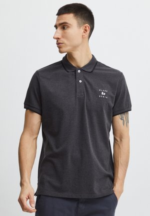 Rövid hajú férfi, aki sötétszürke, kis fehér logóval díszített pólót visel, világos egyszínű háttér előtt áll, balra néz.