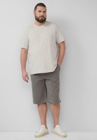 Hellgraues kurzes T-Shirt mit kurzen Ärmeln, kombiniert mit grauen Shorts, die eine lockere Passform haben. Das Modell trägt cremefarbene Sneaker und steht vor einem schlichten Hintergrund.