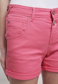 Vaaleanpunaiset farkkushortsit, joissa on käännetty helma. Niissä on kaksi etutaskua ja hopeaiset varusteet vyötäröllä sekä taskujen reunoissa.