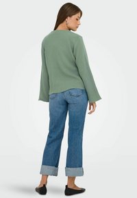 Top largo de manga larga texturizado en verde, combinado con jeans azules claros con puños y bailarinas negras. La blusa tiene un corte relajado con un escote redondo.