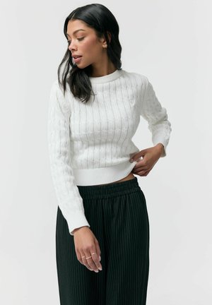 Kobieta o długich ciemnych włosach, ubrana w biały sweter z warkoczowym splotem i czarne spodnie w prążki, pozująca z jedną ręką poprawiającą sweter.