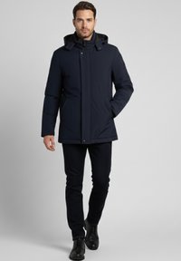 Parka impermeable azul marino con cremallera frontal, capucha desmontable y bolsillos laterales. Combinado con jeans oscuros y zapatos negros.