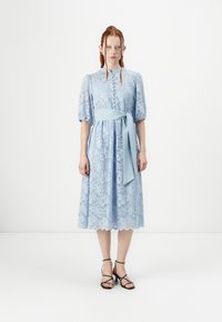 Lyseblå blondekjole med korte puffermer, knapfront og ankerbelte i livet. Bølgeformet kant og blomstermønster. Kombinert med svarte stroppede hæler.