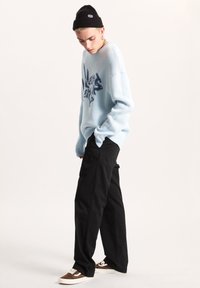 Pullover tricotato azzurro chiaro con un design grafico blu navy, indossato con pantaloni neri a vestibilità ampia e scarpe da ginnastica marroni con lacci. Accessorio berretto di maglina nera.